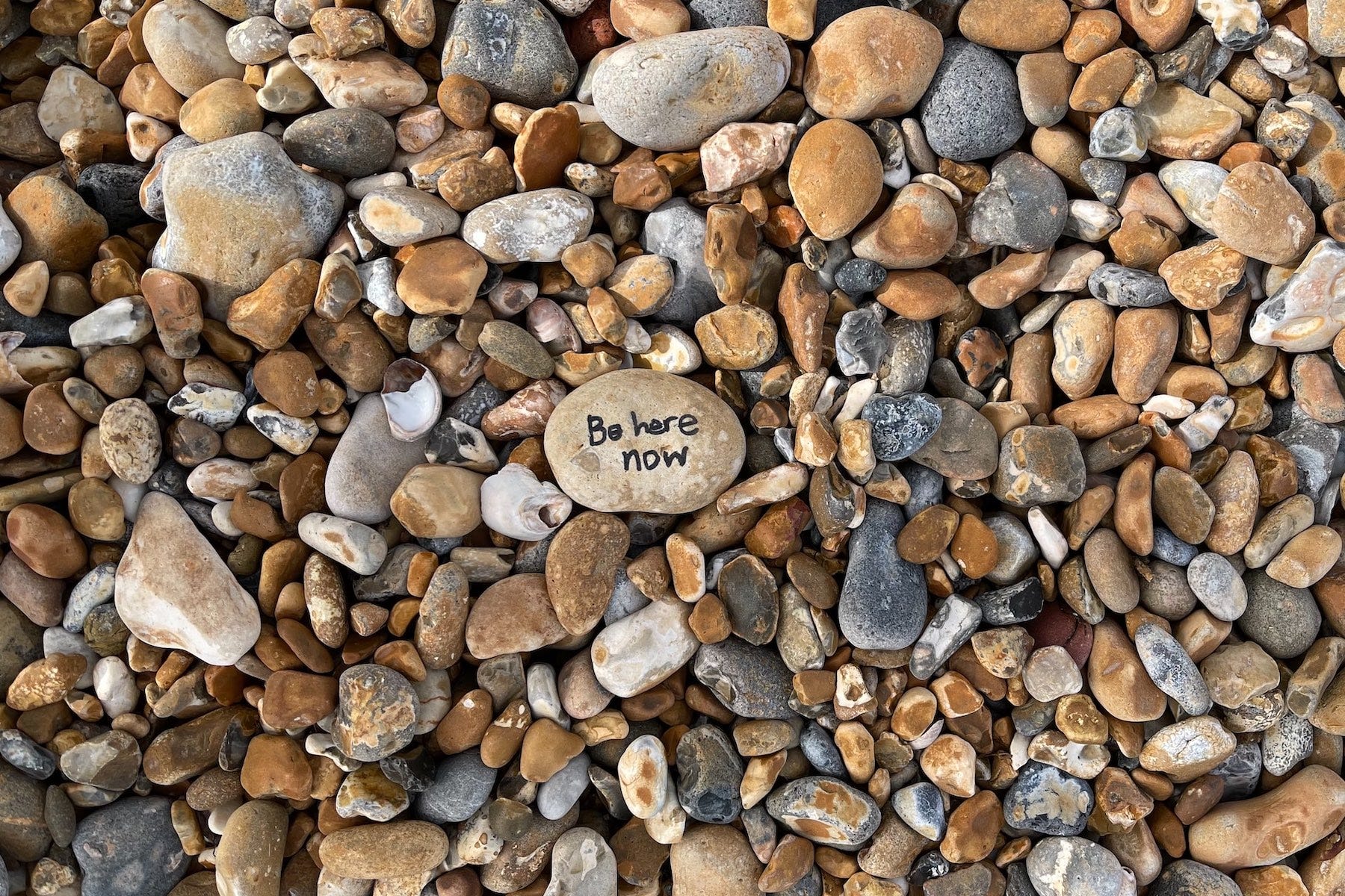 A photo showing rocks of different size. A large rock in the center has Be Here Now written on it.