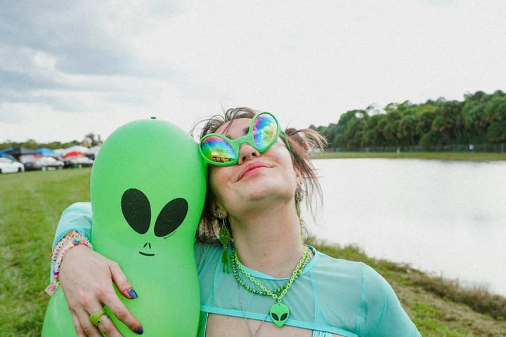 Delight comes in many colors. A person wearing green sunglasses looks up at the sky while holding an inflatable alien.