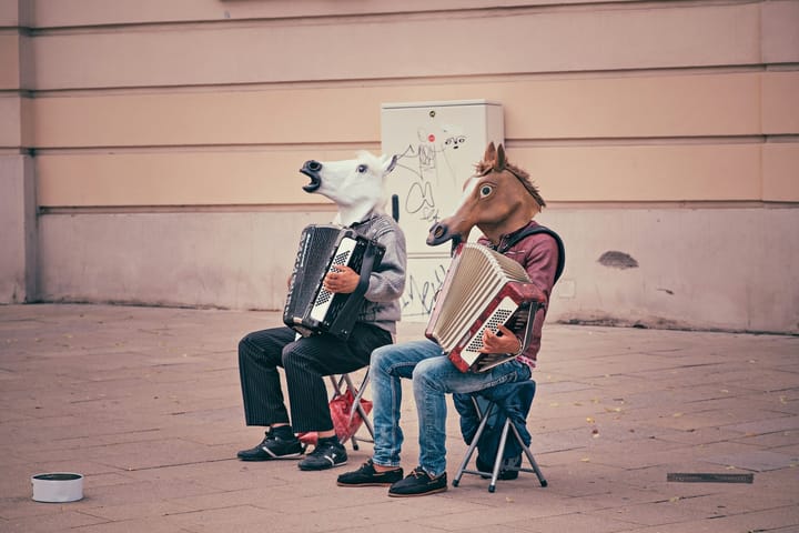 Two people sit on chairs outside a stone building. They are wearing horse heads and playing accordions. 