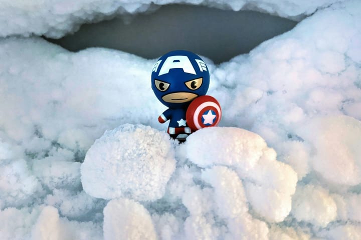 A toy superhero stands holding a shield. It appears that they are in a snowy landscape. 