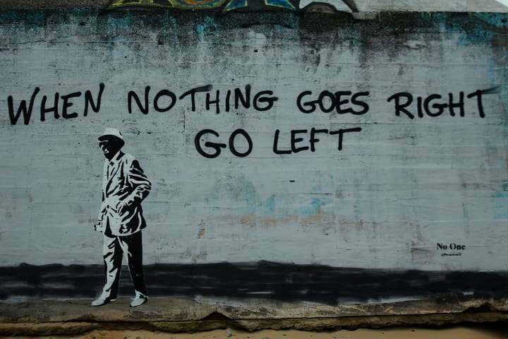 A wall painted with the words When Nothing Goes Right Go Left. A old man in a hat is also painted on the wall. He is walking to the left.